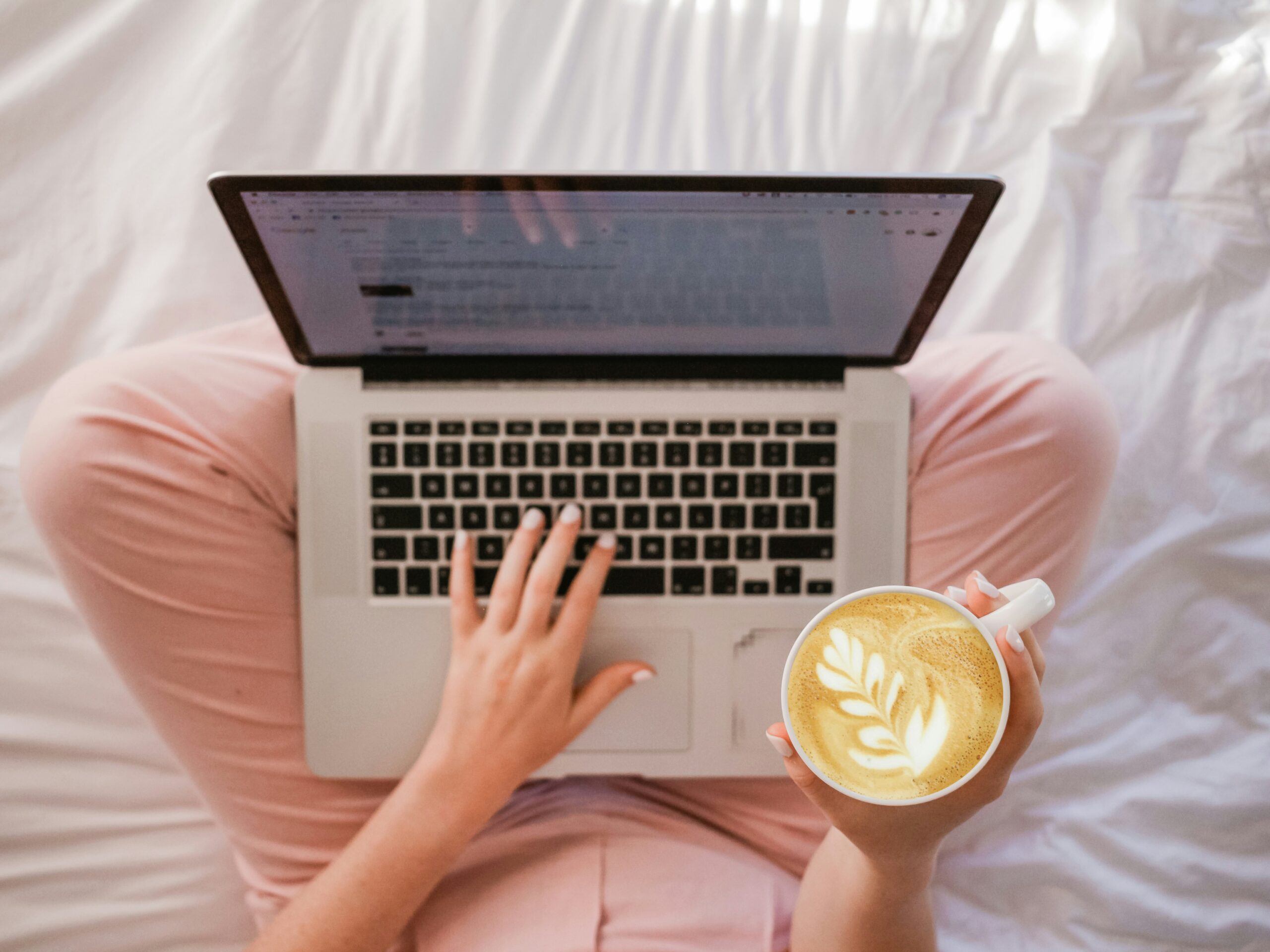 A person sits down cross-legged on their laptop while holding a cup of coffee.